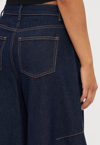 Pantaloni larghi din denim închis, cu cusături portocalii, talie înaltă și două buzunare pe spate. Textură netedă, cu un design clasic.
