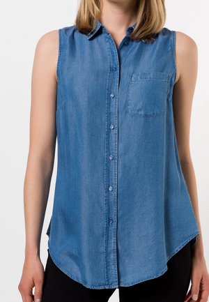 Chemise en denim sans manches de couleur bleu clair, avec un col, des boutons à l'avant, une poche poitrine et un ourlet arrondi. Le tissu est lisse et texturé.