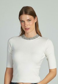 Top bianco a maniche corte con scollatura rotonda decorata con un'applicazione di perle argentate, caratterizzato da una texture liscia e una vestibilità aderente.