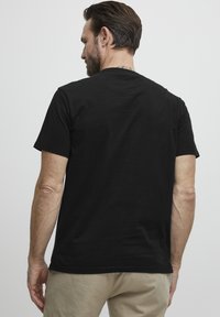 Juodos medvilnės marškinėliai, trumpomis rankovėmis, apvaliu kaklu, vientisas dizainas, be raštų ar akcentų, glotni tekstūra, dėvimi su smėlio spalvos kelnių.