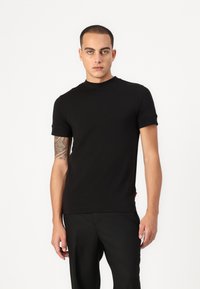 Černé tričko s krátkým rukávem a kulatým výstřihem, vyrobené z hladké látky. Model je nosí s tailored černými kalhotami.