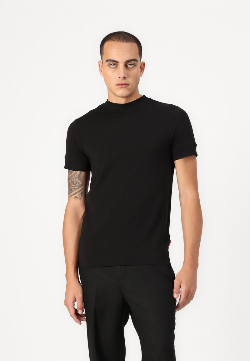 Černé tričko s krátkým rukávem a kulatým výstřihem, vyrobené z hladké látky. Model je nosí s tailored černými kalhotami.