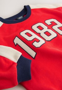 Sweat-shirt rouge avec le chiffre "1982" en crème avec contour marine. Manches contrastantes marine et crème, poignets et col côtelés.