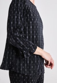 Schwarze plissierte Bluse mit dezenten weißen Mustern, langen Ärmeln und Rundhalsausschnitt, mit gerüschten Bündchen und einem lockeren Schnitt.