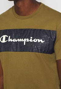 Muž v olivově zeleném tričku s černým vodorovným panely, na kterém se přes hruď nachází bílé logo značky Champion.