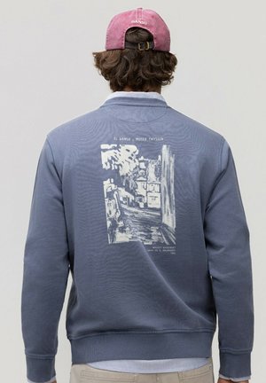 Persona que lleva una sudadera azul con un estampado blanco de paisaje urbano y texto, combinada con una gorra de béisbol rosa puesta al revés.