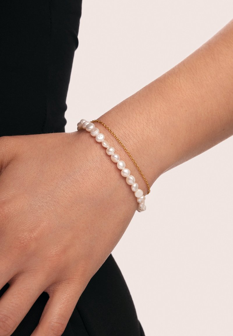 Dos pulseras en una muñeca: una con perlas blancas y otra de cadena dorada. La textura de la piel es visible y el fondo es neutral.