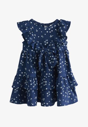 Lächelndes junges Mädchen mit hellbraunen Haaren, das ein blaues Kleid mit kleinen weißen Blumen und einer großen Schleife trägt, steht vor einer hellen Wand.