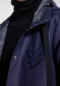 Personne portant une veste bleu marine avec un emblème d'oiseau noir sur un pull à col roulé côtelé noir.