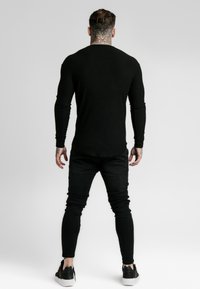 Schwarzes, geripptes Langarmshirt kombiniert mit schwarzen Skinny Jeans und schwarzen Sneakers, mit abgerundetem Saum und figurbetontem Schnitt, der Tätowierungsdetails zur Geltung bringt.