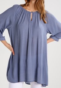 Blusa tunica a maniche lunghe di colore azzurro con scollatura a chiave e dettaglio plissettato. Tessuto morbido e testurizzato con vestibilità ampia e orlo arrotondato.