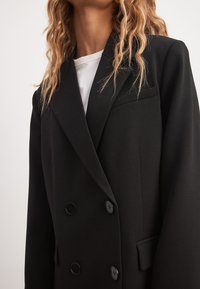 Musta kaksirivinen bleiseri sileästä kankaasta, jossa on huippukaulukset, kaksi etunappia ja rintatasku. Ei näkyviä kuvioita tai yksityiskohtia.