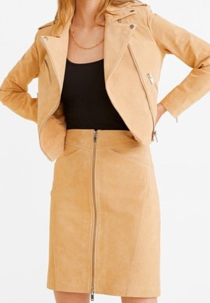 Femme portant une veste motard en daim beige et une jupe assortie à fermeture éclair sur un haut noir, debout les mains sur les hanches devant un fond blanc.