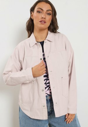 Vrouw draagt een lichtroze button-up jas over een zwart shirt met print en blauwe spijkerbroek, staand tegen een effen witte achtergrond.