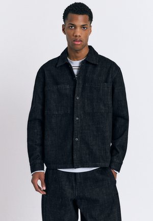 Giovane uomo con giacca e pantaloni di jeans neri, in piedi davanti a uno sfondo chiaro e uniforme, rivolto in avanti con espressione neutra.