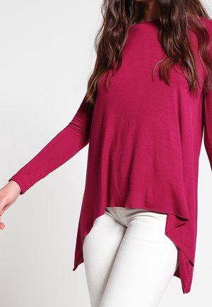 Frau trägt ein asymmetrisches, langärmliges magentafarbenes Oberteil mit eng anliegenden weißen Hosen, gezeigt von den Schultern bis zur Hüfte vor schlichtem Hintergrund.