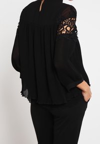 Czarna plisowana bluzka z przezroczystymi koronkowymi akcentami na rękawach, wysokim dekoltem i luźnym krojem. Miękki, lekki materiał z płynącą sylwetką.