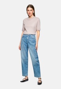 Helles graues kurzärmliges Oberteil, hoch-taillierte hellblaue Jeans und schwarze flache Schuhe. Das Model steht mit den Händen an den Seiten.