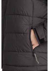 Čierna prešívaná bunda s prešívaným vzorom, so zapínaním na zips a vreckom na zips, ktoré sa zaviera na suchý zips. Obsahuje textúrovanú pútko na zips.