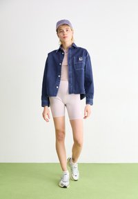 Cămașă din denim de culoare albastru închis, cu închidere prin butoane și buzunar pe piept, asortată cu un body de culoare bej deschis și pantaloni scurți de bicicletă asortați, completată cu adidași argintii.