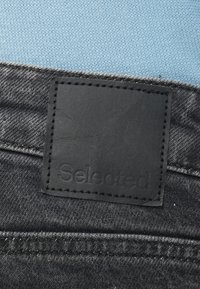 Remendo de couro preto com a palavra "Selected" em relevo em denim cinza. O tecido apresenta uma leve textura com costura visível ao longo das bordas.