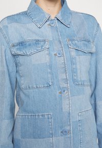 Světle modrá denimová bunda s patchworkovým designem, dvěma náprsními kapsami a modrými knoflíky. Má límec a klasický zapínací styl na knoflíky.