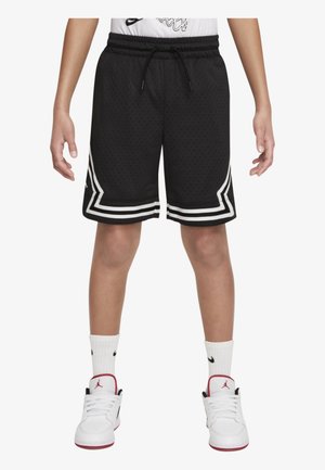 Kind draagt zwarte basketbalshorts met witte bies, witte broeksokken en witte sneakers met rode accenten, staat met gespreide benen.