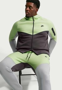 Homme assis sur une chaise portant un survêtement Nike color-block vert, gris et noir avec un sweat à capuche zippé et un pantalon assorti.