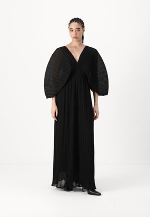 Black pleated maxi dress with wide, draped sleeves, deep V-neckline, smooth texture, and flowing silhouette, paired with black heeled boots.