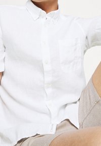 Camicia bianca a maniche corte con bottoni, realizzata in lino, con taschino sul petto, colletto casual e un'etichetta visibile all'orlo.