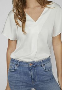 Blusa blanca de manga corta con escote en V, hecha de tela suave; combinada con unos vaqueros de mezclilla azul claro que tienen botón y bolsillos.