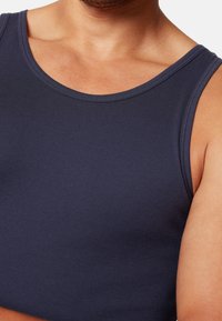 Granatowy top na ramiączkach wykonany z gładkiego materiału, z okrągłym dekoltem i szerokimi wycięciami na ramionach, podkreślający dopasowany krój w okolicy torsu.