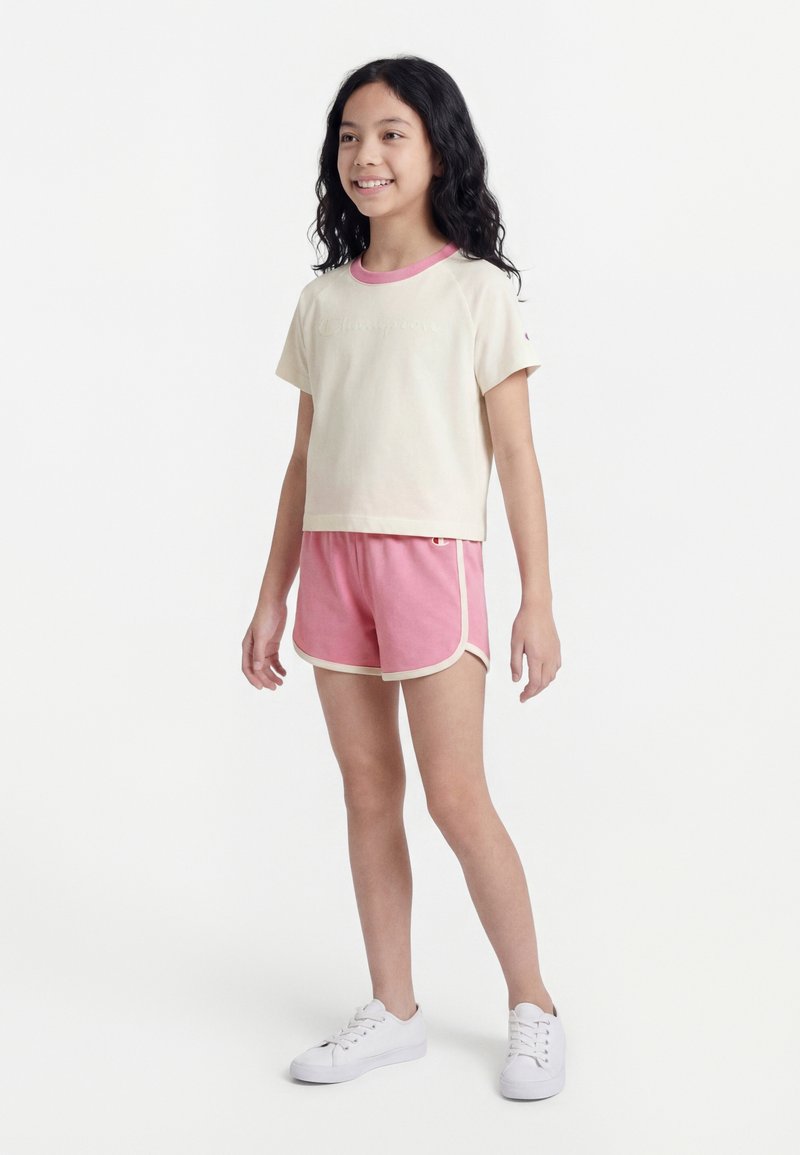 Jeune fille debout, portant un t-shirt à manches courtes couleur crème avec un col rose et un short rose avec un bord crème, chaussures blanches sur fond blanc.