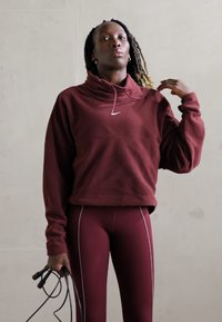 Bordeauxfarbener Sweatshirt mit hohem Kragen und weißem Logo, kombiniert mit passenden Leggings mit kontrastierenden weißen Nähten, gehalten mit einem Springseil.