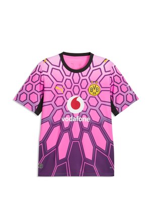 Jeune homme portant une casquette noire Puma, une veste noire Puma, un maillot de football violet du Borussia Dortmund, un pantalon noir et des baskets noir et blanc, debout devant un fond gris.