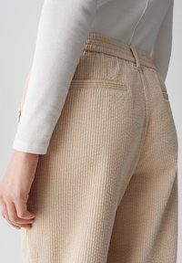 Beige Cordhosen mit vertikalen Rippungen, mit hoher Taille, Gesäßtaschen und einem lockeren Schnitt. Getragen mit einem weißen Langarmoberteil.