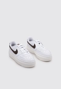 Zapatillas deportivas de cuero blanco con textura suave, punta redonda, perforaciones en la puntera y logotipo de Nike Swoosh en contraste de color marrón oscuro a los lados.