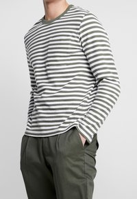 Langärmliges Hemd mit grünen und weißen horizontalen Streifen, ausgestattet mit einer Brusttasche und Rundhalsausschnitt, kombiniert mit grünen Hosen.