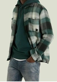 Grön- och svartmönstrad flanellskjorta med vävd struktur, över en mörkgrön huvtröja; kombinerad med ljusblå jeans.