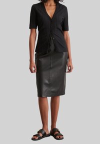 Blusa corta de manga corta negra con canalé, escote en V y botones frontales, combinada con una falda midi ajustada de cuero negra y sandalias planas negras.