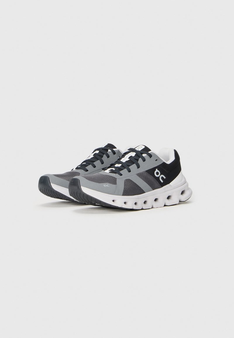 Graue Sportschuhe mit einem Mesh-Obermaterial, schwarzen Akzenten, weißer Sohle und strukturierten Grip-Mustern. Mit Schnürsenkeln und Logodetails an der Seite.