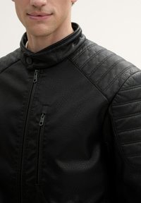 Giubbotto in pelle nera con colletto alto, chiusura con zip e pannelli testurizzati sulle spalle. Il materiale ha una finitura a granulato.