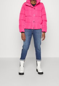 Person som bär en klar pink puffjacka, upprullade blå jeans och vita ankellång stövlar står mot en enkel ljus bakgrund.