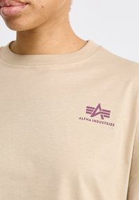 Kreminės spalvos medvilninė marškinėliai su mažuoju violetiniu "Alpha Industries" logotipu kairėje krūtinės pusėje. Apvalus iškirpimas ir trumpomis rankovėmis. Lygus paviršius.