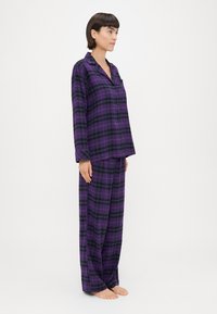 Μωβ και μαυρόκουμπα πιτζάμα, με μπλούζα μακρυμάνικη και κουμπιά, και χαλαρούς παντελόνες, φτιαγμένη από μαλακό ύφασμα.