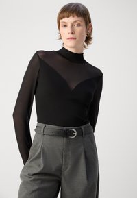 Top nero a maniche lunghe con pannello superiore in rete trasparente, abbinato a pantaloni grigi a vita alta e una cintura nera con cinturino in argento.