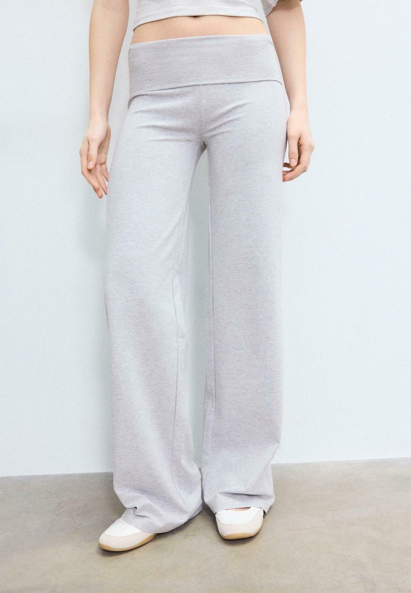 Parte inferiore del corpo di una persona che indossa pantaloni della tuta larghi e grigio chiaro e scarpe bianche senza lacci, in piedi contro un muro di colore chiaro.