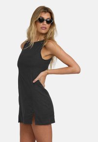 Black sleeveless dress with side slits, fitted design, smooth texture, and mixed materials. Model wears black cat-eye sunglasses.