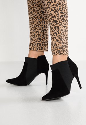 Černé semišové kotníkové boty s špičatou špičkou, vysokým jehlovým podpatkem a elastickými bočními panely. Nosené s kalhotami s leopardím potiskem.