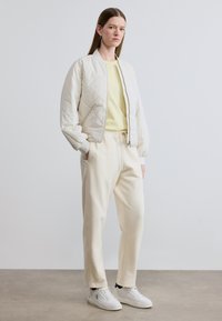 Εκρού μπουφάν bomber με ραφές πάνω σε ανοιχτό κίτρινο πουκάμισο, συνδυασμένο με λευκό παντελόνι με κορδόνια και λευκά αθλητικά παπούτσια. Το δάπεδο είναι γκρι σκυρόδεμα.